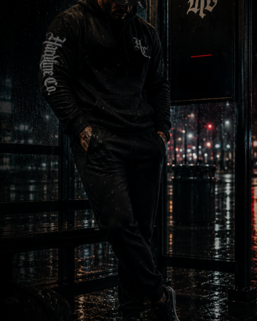 Person wearing a black hoodie and pants standing in a dark, urban setting with neon signs.