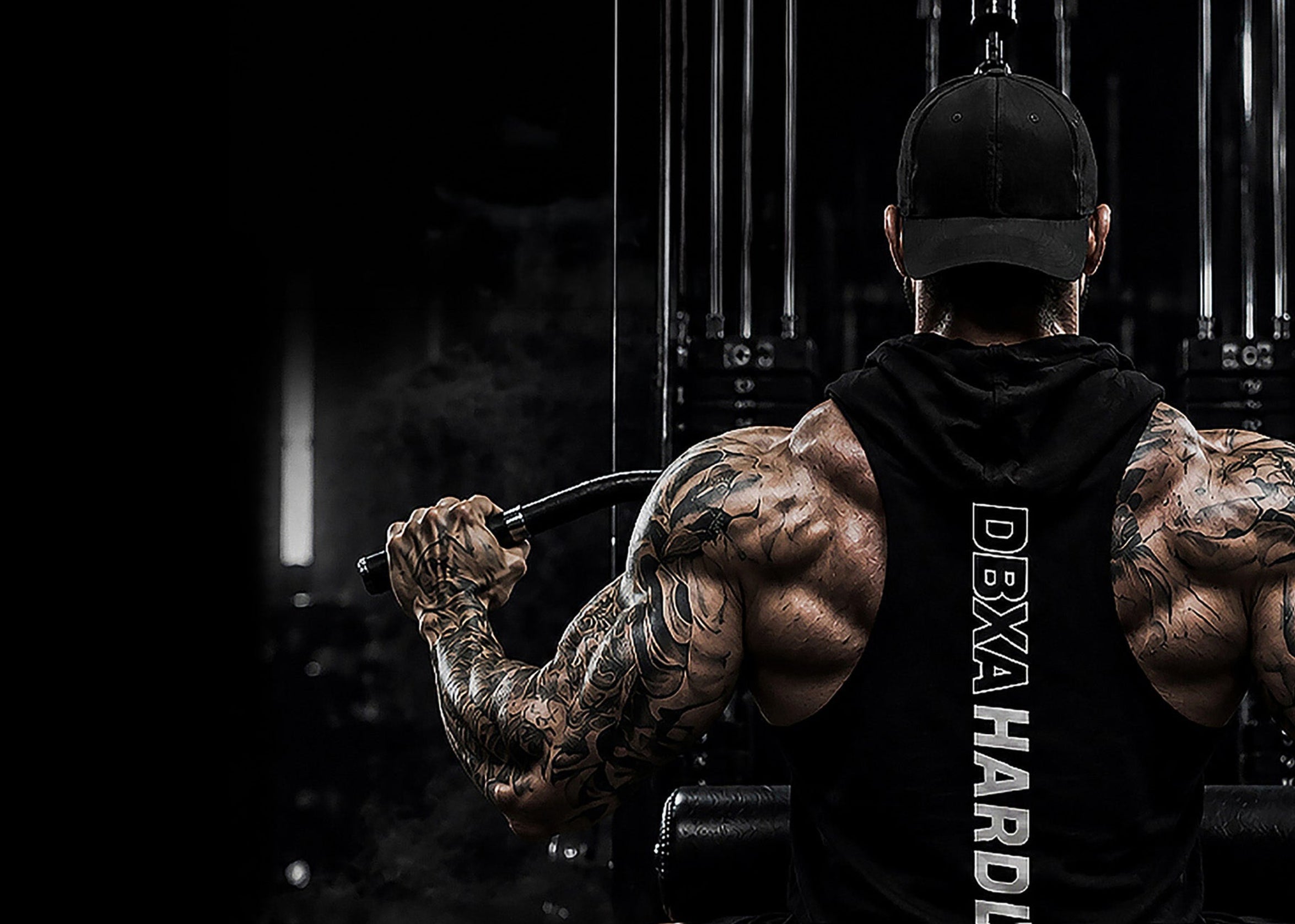 Muscular man with tattoos lifting weights in a gym setting, wearing a black 'Dexa Hardline' hoodie.