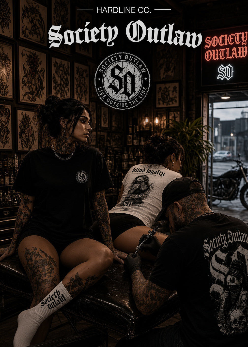 Three people in a tattoo studio wearing Society Outlaw apparel, with one person seated on a table and another receiving a tattoo. The scene includes visible Society Outlaw logos and branding on clothing, wall signage, and a motorcycle outside the window.