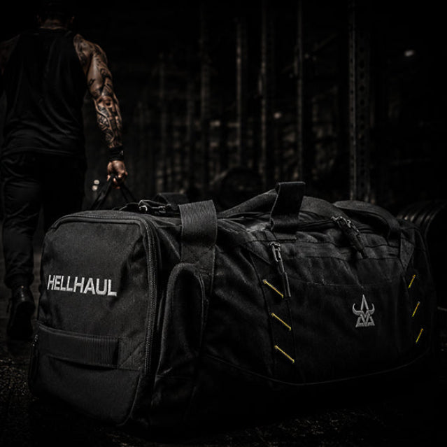 Person walking with a large black duffel bag labeled 'HELLHAUL' in a dark setting.