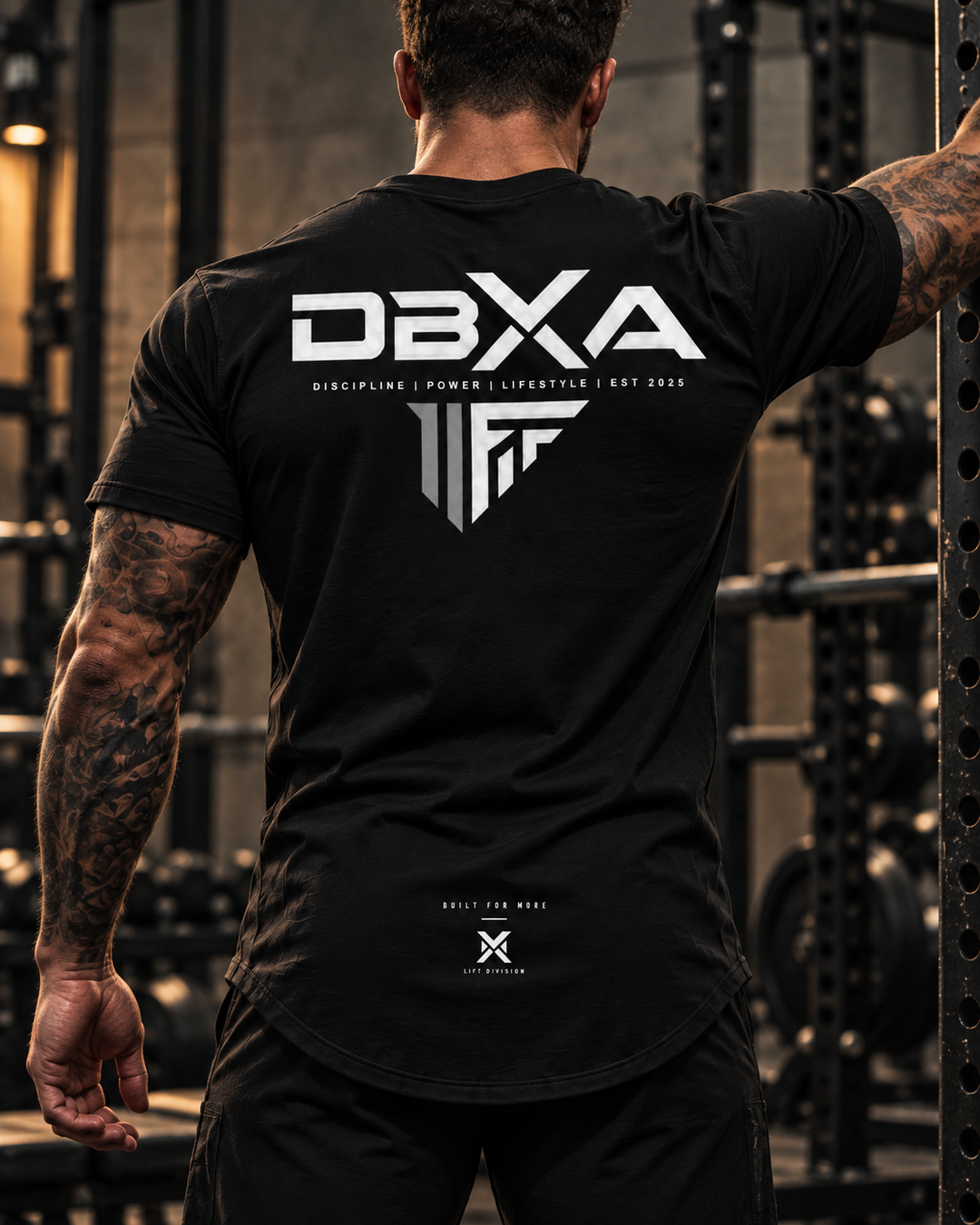 Tattooed male model wearing the black DBXA Stronghold Tee, rear view in an industrial gym.