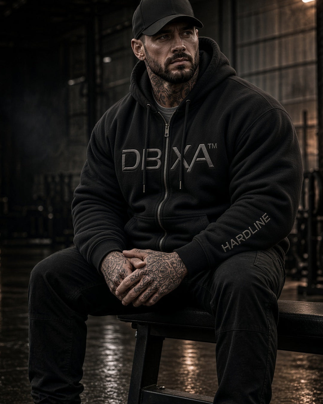 A man wearing a black DBXA hoodie and matching sweatpants sits on a bench in a dark gym environment. The hoodie features a large 'DBXA' logo on the chest and 'HARROULE' on the left sleeve.