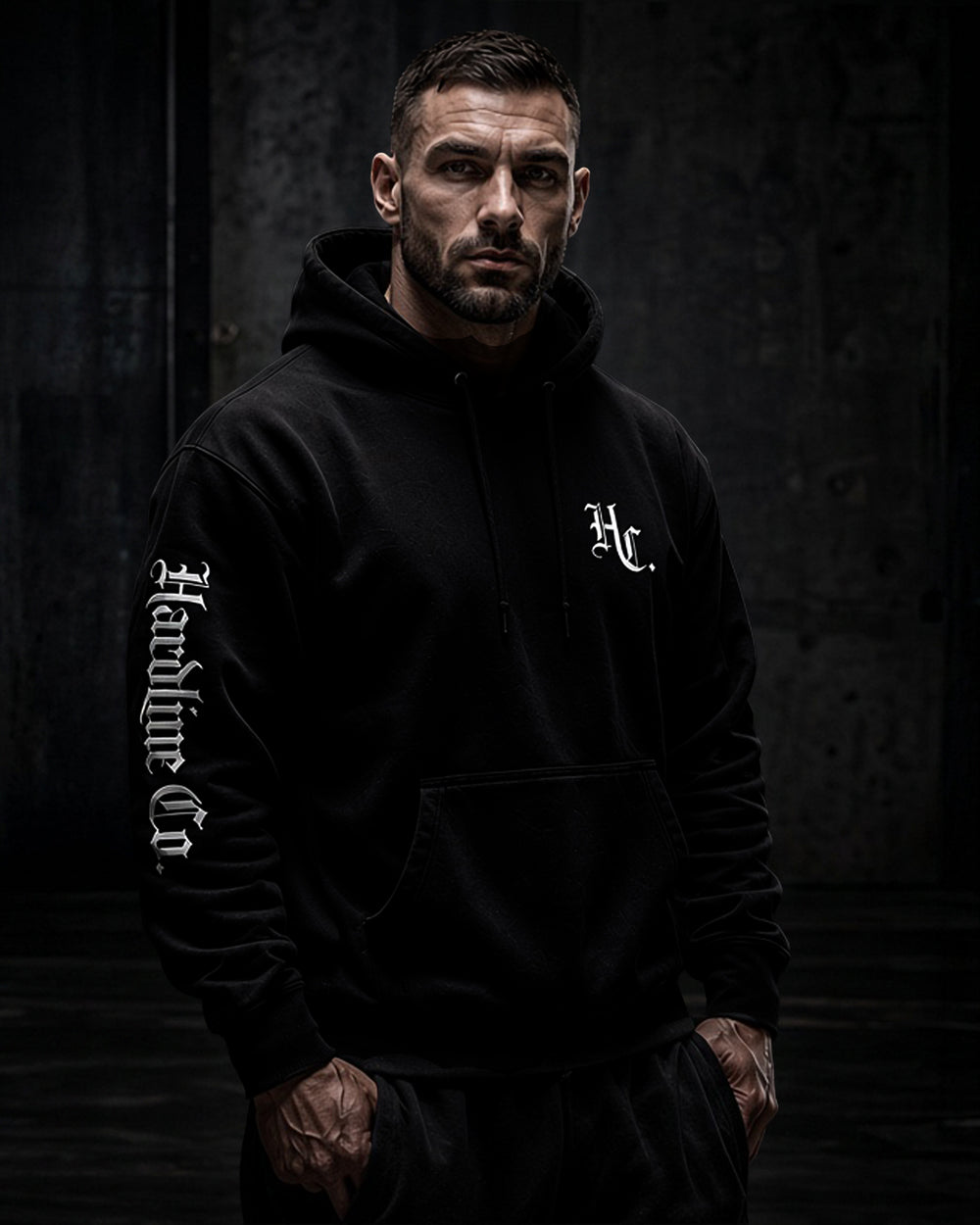 A man wearing a black pullover hoodie with white gothic-style text on the left sleeve and a white monogram on the left chest, posed against a dark textured studio background.