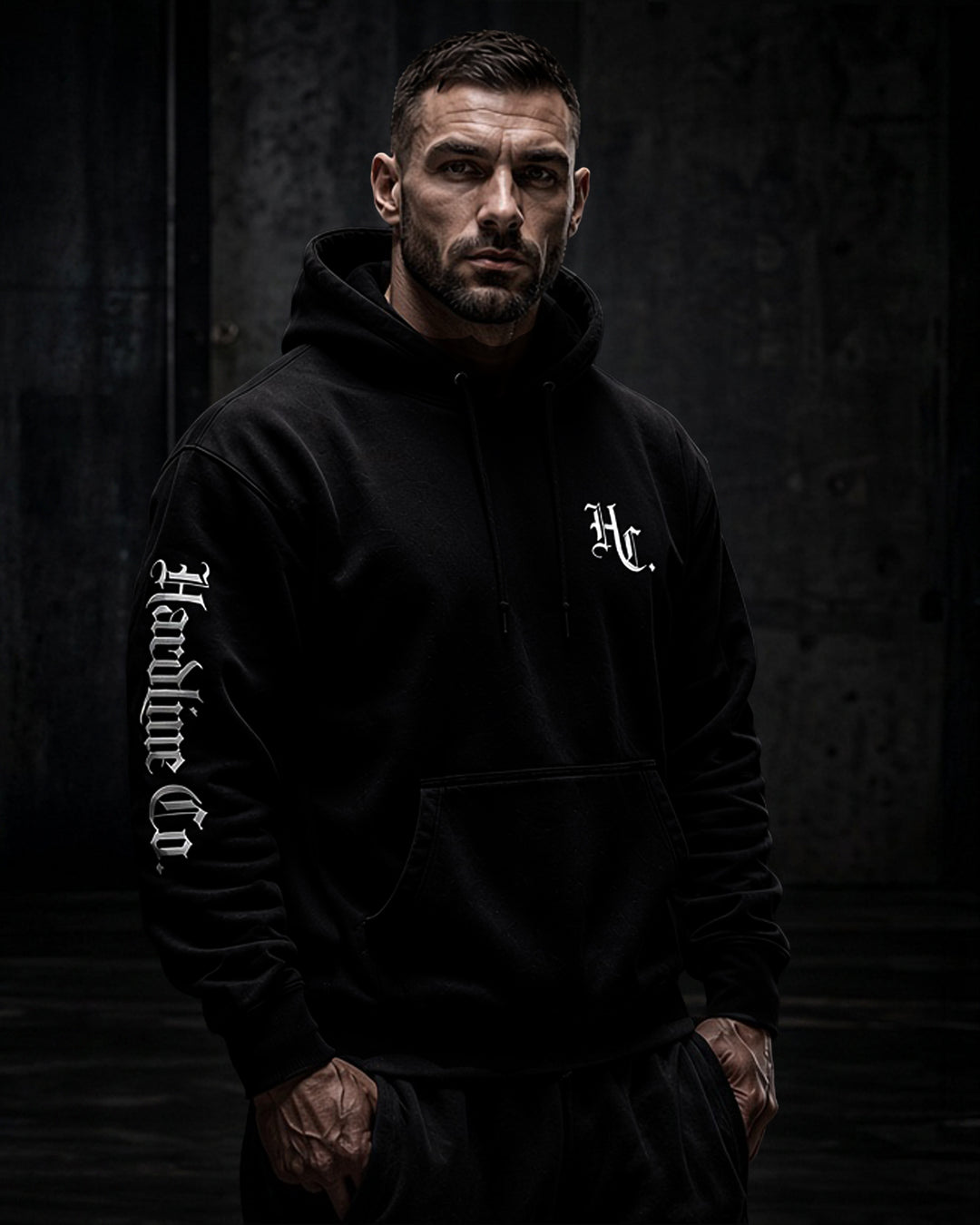 A man wearing a black pullover hoodie with white gothic-style text on the left sleeve and a white monogram on the left chest, posed against a dark textured studio background.