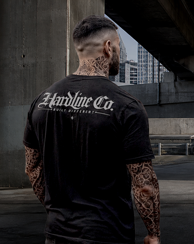A man with tattoos is shown from behind wearing a black t-shirt with 'Hardline Co. Built Different' printed in white across the back, standing in an urban concrete environment.