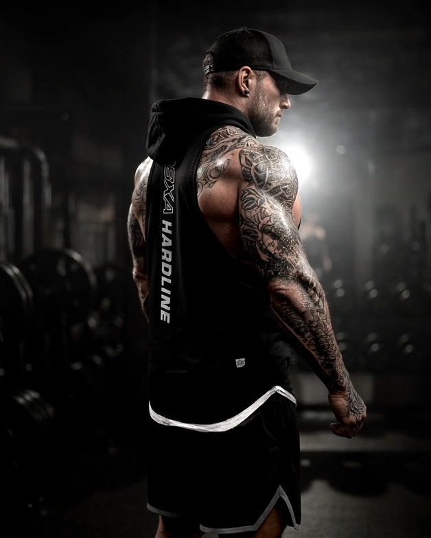 Man with tattoos in a gym setting wearing a black tank top with text.