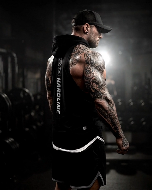 Man with tattoos in a gym setting wearing a black tank top with text.