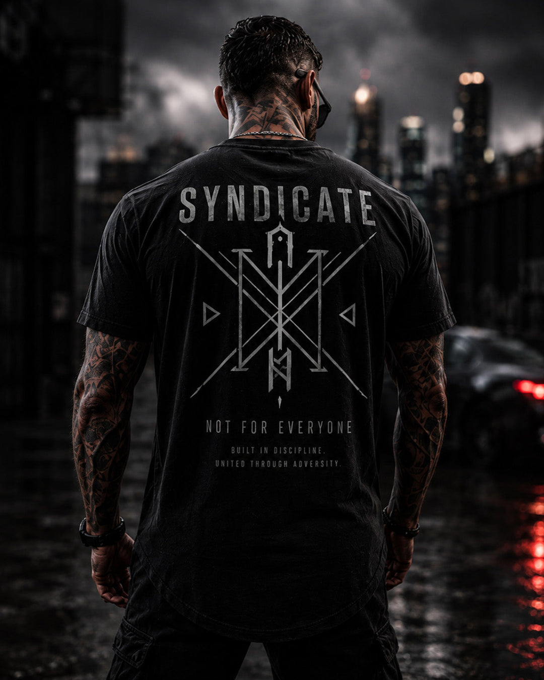 A man viewed from behind wearing a black t-shirt with a large white graphic and the word 'SYNDICATE' printed on the back, standing on a wet city street at night.