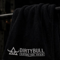 Black towel with 'DIRTYBULL' logo hanging on a barbell in a gym setting